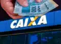 Mão segurando notas de real em frente à fachada de uma agência da Caixa Econômica Federal, representando o Saque Calamidade do FGTS de até R$ 6.220 para moradores de municípios atingidos por desastres naturais