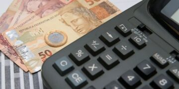Calculadora financeira preta sobre uma mesa, ao lado de uma moeda de 1 Real e notas de 50 Reais, ilustrando o cálculo de impostos e planejamento financeiro.