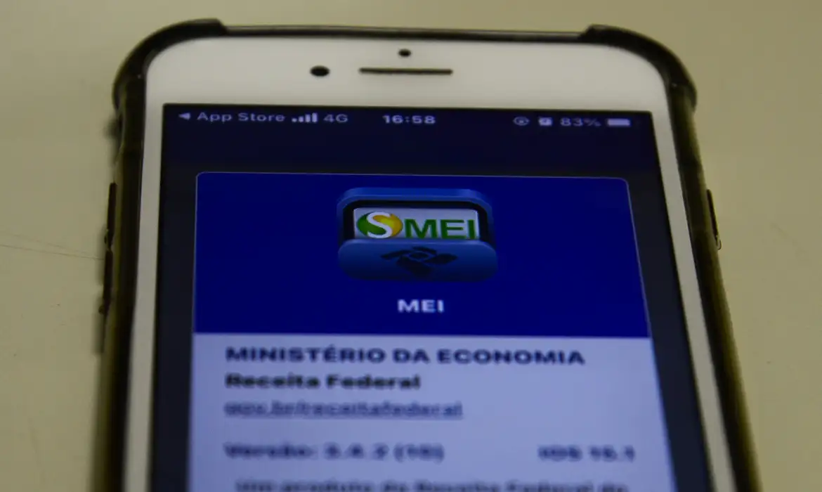 Visão aproximada de um celular iPhone exibindo o ícone azul do aplicativo "SMEI" da Receita Federal. O dispositivo está sobre uma mesa clara, mostrando detalhes da interface do Ministério da Economia.