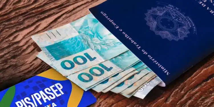 Uma Carteira de Trabalho azul aberta sobre uma mesa de madeira, com várias cédulas de 100 reais saindo de dentro dela. Ao lado, um cartão azul e amarelo com a inscrição "PIS/PASEP CAIXA".