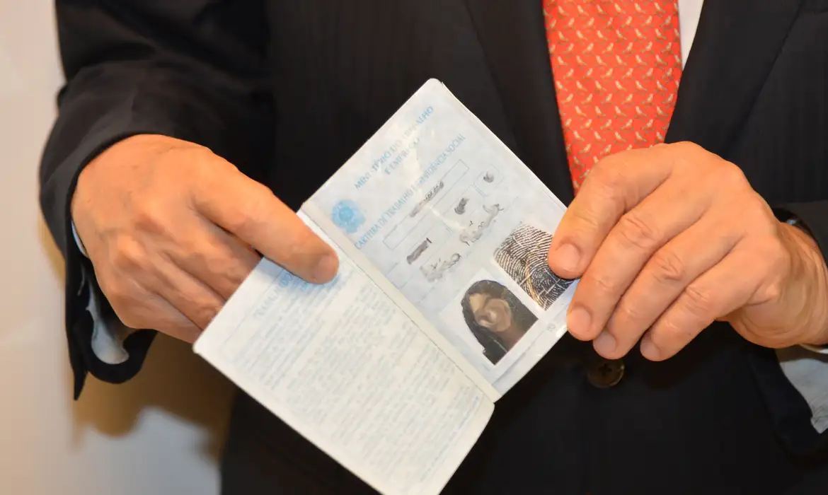 quem tem direito seguro desemprego 2026 regras Close-up de mãos segurando uma Carteira de Trabalho (CTPS) aberta, evidenciando a página de identificação, em frente a uma pessoa vestindo terno e gravata.