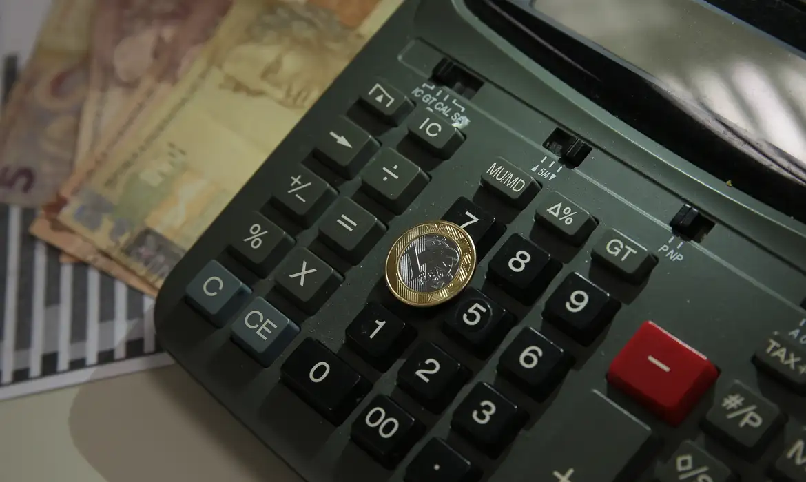 Calculadora com moeda brasileira sobre as teclas Calculadora preta com moeda de real posicionada sobre as teclas e cédulas ao fundo