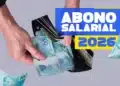 Mãos tiram notas de cem reais de uma carteira preta com destaque para texto "Abono Salarial 2026".