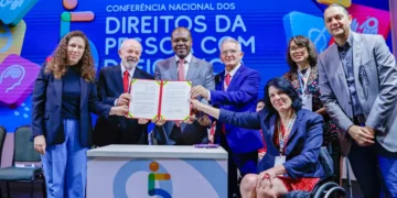 Grupo de sete pessoas, incluindo mulher em cadeira de rodas, segura documento aberto em evento oficial