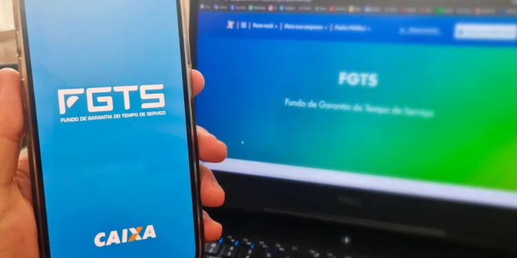 Mão segura celular com app FGTS aberto próximo a notebook exibindo site do FGTS ao fundo
