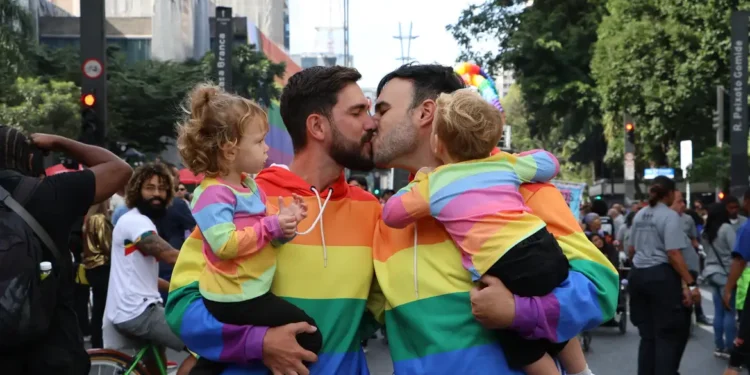 Dois homens se beijam segurando crianças nos braços, todos com roupas de arco-íris durante evento de rua