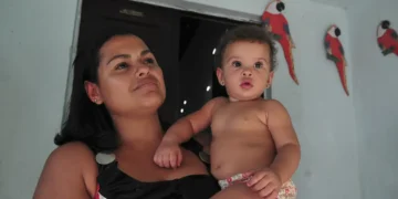 Mulher segura bebê no colo em frente a parede azul com araras de madeira penduradas