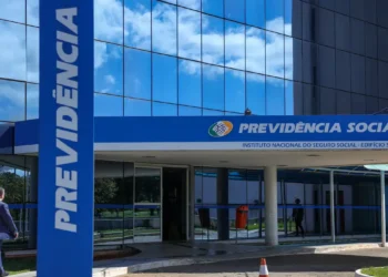 Entrada principal da Previdência Social com vidro refletindo céu azul e pessoa caminhando
