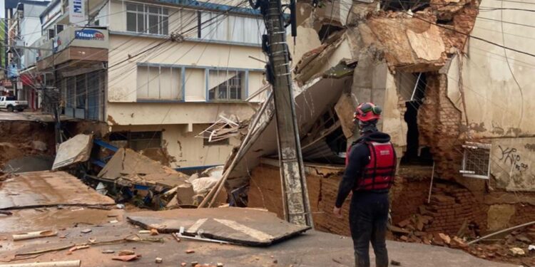 Imagem de um prédio desmoronado devido às chuvas em Minas Gerais, com um socorrista observando a cena de destruição, incluindo fios caídos e detritos espalhados pela rua.