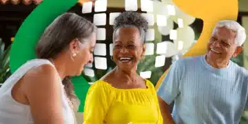 Três idosos sorrindo sentados juntos com decoração colorida ao fundo.