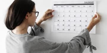 Mulher de óculos prende calendário na parede enquanto observa datas marcadas