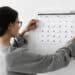 Mulher de óculos prende calendário na parede enquanto observa datas marcadas