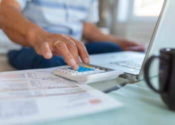 Mão digitando em calculadora ao lado de documentos e notebook