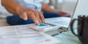 Mão digitando em calculadora ao lado de documentos e notebook