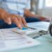 Mão digitando em calculadora ao lado de documentos e notebook