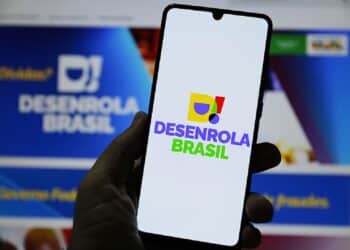 Mão segurando smartphone com logotipo colorido do Desenrola Brasil na tela, fundo desfocado do site gov.br