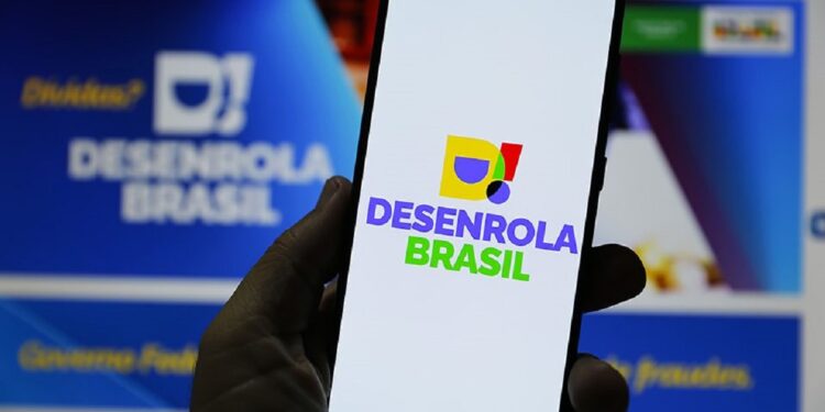 Mão segurando smartphone com logotipo colorido do Desenrola Brasil na tela, fundo desfocado do site gov.br