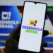 Mão segurando smartphone com logotipo colorido do Desenrola Brasil na tela, fundo desfocado do site gov.br