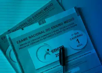 Close de dois cadernos de prova oficiais do Enem (um rosa e um azul) empilhados, sob uma iluminação azul ciano. Uma caneta preta está posicionada na parte inferior do caderno azul, que exibe "1º DIA - CADERNO 1 AZUL".