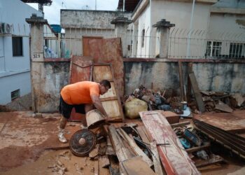 Homem de camiseta laranja remove móveis e objetos destruídos pela lama de enchente em área residencial coberta por barro.