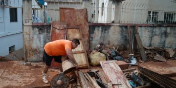 Homem de camiseta laranja remove móveis e objetos destruídos pela lama de enchente em área residencial coberta por barro.