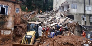 Bombeiros e equipes de resgate trabalham em escombros de casas destruídas por deslizamento, com retroescavadeira CAT amarela.