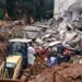 Bombeiros e equipes de resgate trabalham em escombros de casas destruídas por deslizamento, com retroescavadeira CAT amarela.