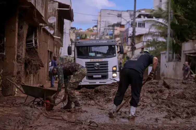 Trabalhadores removem lama de rua com enxadas e pás enquanto caminhão Constellation aguarda em área atingida por enchente.