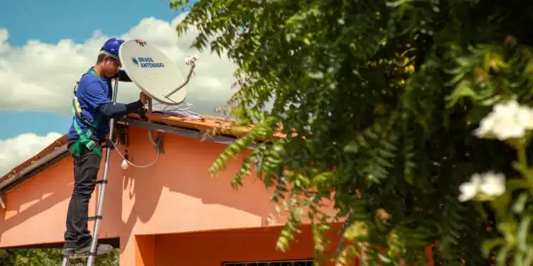 Técnico em escada instala antena parabólica digital Brasil Antenado em telhado de casa laranja.