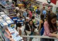 Vista aérea do interior de um supermercado ou loja de departamentos movimentada. Pessoas circulam pelos corredores com cestas de compras e aguardam em filas nos caixas, cercadas por prateleiras repletas de produtos.