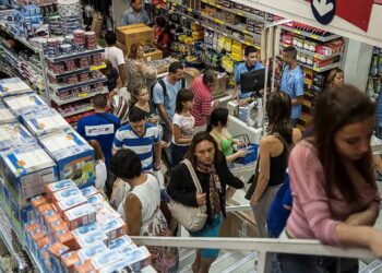 Vista aérea do interior de um supermercado ou loja de departamentos movimentada. Pessoas circulam pelos corredores com cestas de compras e aguardam em filas nos caixas, cercadas por prateleiras repletas de produtos.