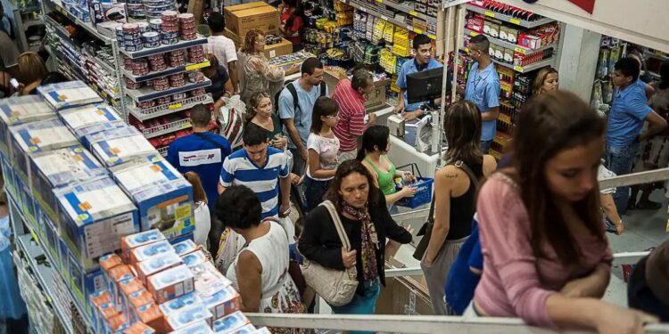 Vista aérea do interior de um supermercado ou loja de departamentos movimentada. Pessoas circulam pelos corredores com cestas de compras e aguardam em filas nos caixas, cercadas por prateleiras repletas de produtos.