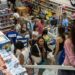 Vista aérea do interior de um supermercado ou loja de departamentos movimentada. Pessoas circulam pelos corredores com cestas de compras e aguardam em filas nos caixas, cercadas por prateleiras repletas de produtos.