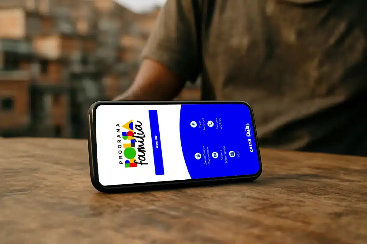 Celular exibindo aplicativo do Bolsa Família sobre mesa de madeira com fundo urbano desfocado