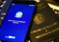 Um smartphone exibindo o aplicativo da Carteira de Trabalho Digital com o brasão da República sobre uma tela azul. O aparelho está posicionado em cima de uma Carteira de Trabalho e Previdência Social (CTPS) física de cor preta com letras douradas.