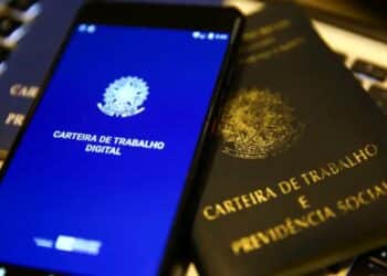 Um smartphone exibindo o aplicativo da Carteira de Trabalho Digital com o brasão da República sobre uma tela azul. O aparelho está posicionado em cima de uma Carteira de Trabalho e Previdência Social (CTPS) física de cor preta com letras douradas.