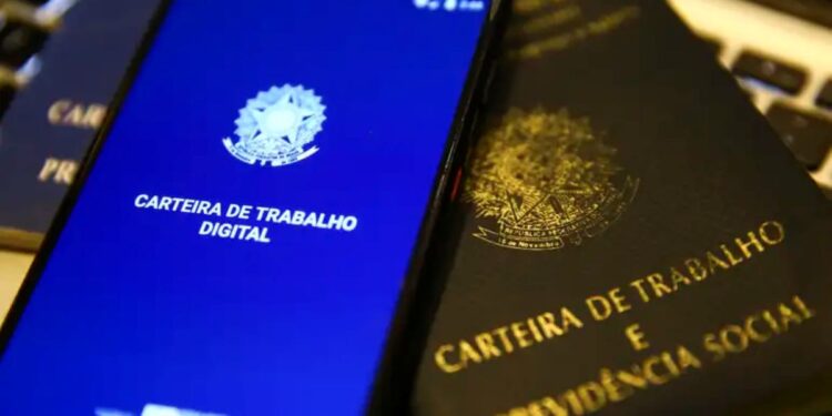 Um smartphone exibindo o aplicativo da Carteira de Trabalho Digital com o brasão da República sobre uma tela azul. O aparelho está posicionado em cima de uma Carteira de Trabalho e Previdência Social (CTPS) física de cor preta com letras douradas.