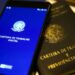Um smartphone exibindo o aplicativo da Carteira de Trabalho Digital com o brasão da República sobre uma tela azul. O aparelho está posicionado em cima de uma Carteira de Trabalho e Previdência Social (CTPS) física de cor preta com letras douradas.