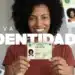 Uma mulher negra sorridente, com cabelos cacheados, segurando com as duas mãos o novo cartão da Carteira de Identidade Nacional (CIN). Ao fundo, há elementos gráficos do documento e o texto "NOVA IDENTIDADE" em destaque.