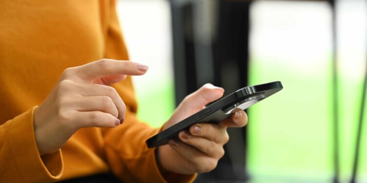 Mãos femininas usando smartphone preto, pessoa com blusa laranja tocando tela do celular em ambiente interno