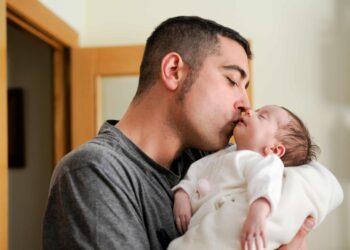 Homem segura e beija um bebê dormindo no colo, próximo a uma porta de madeira