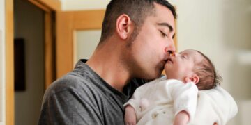 Homem segura e beija um bebê dormindo no colo, próximo a uma porta de madeira