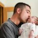 Homem segura e beija um bebê dormindo no colo, próximo a uma porta de madeira