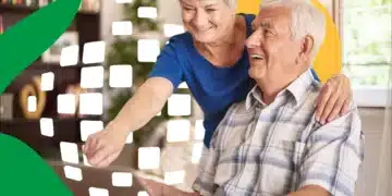 Casal idoso sorri enquanto mulher de blusa azul aponta para tela do computador, homem de camisa xadrez observa atento.