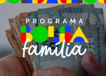 Mão segurando notas de R$ 100 com logo do Programa Bolsa Família em cores verde, amarelo, azul e vermelho.