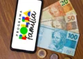 Celular exibindo logo do Bolsa Família sobre mesa de madeira com notas de R$ 20, R$ 50 e R$ 100 e moedas.