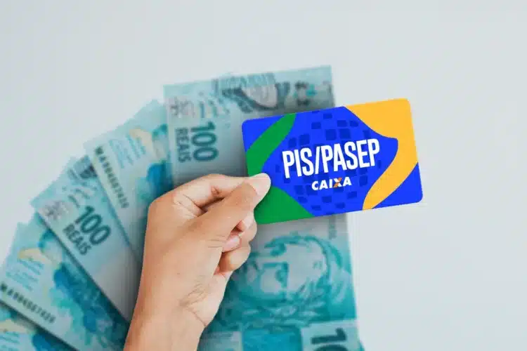 Mão segura cartão PIS/PASEP com logo da Caixa em azul, verde e amarelo sobre notas de 100 reais.