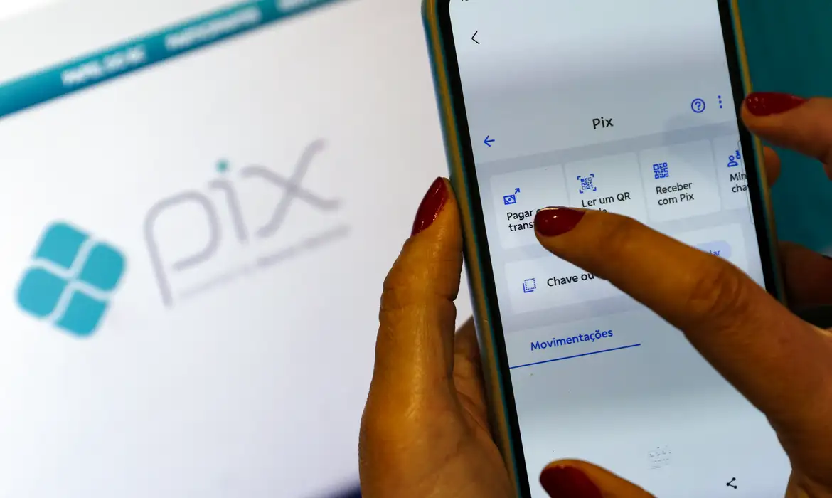 Mãos de uma pessoa com unhas pintadas de vermelho manuseando um smartphone, acessando as opções de "Pagamentos" e "Transferências" no aplicativo do Pix. Ao fundo, uma tela desfocada exibe o logo oficial do Pix.