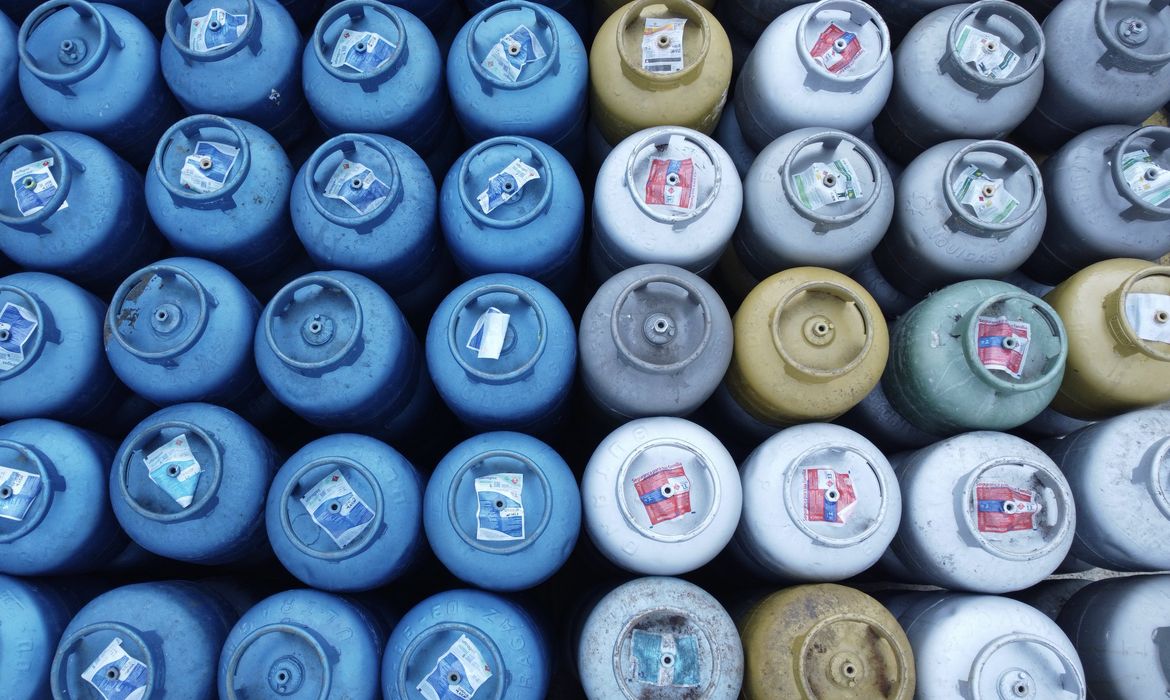 Fileiras de botijões de gás de diferentes cores — azul, branco, amarelo e verde — organizados lado a lado em uma revendedora credenciada pelo programa Gás do Povo