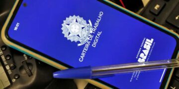 Um smartphone sobre um teclado de computador exibindo a tela azul do aplicativo Carteira de Trabalho Digital do Governo Federal do Brasil. Uma caneta esferográfica azul está pousada sobre o aparelho.